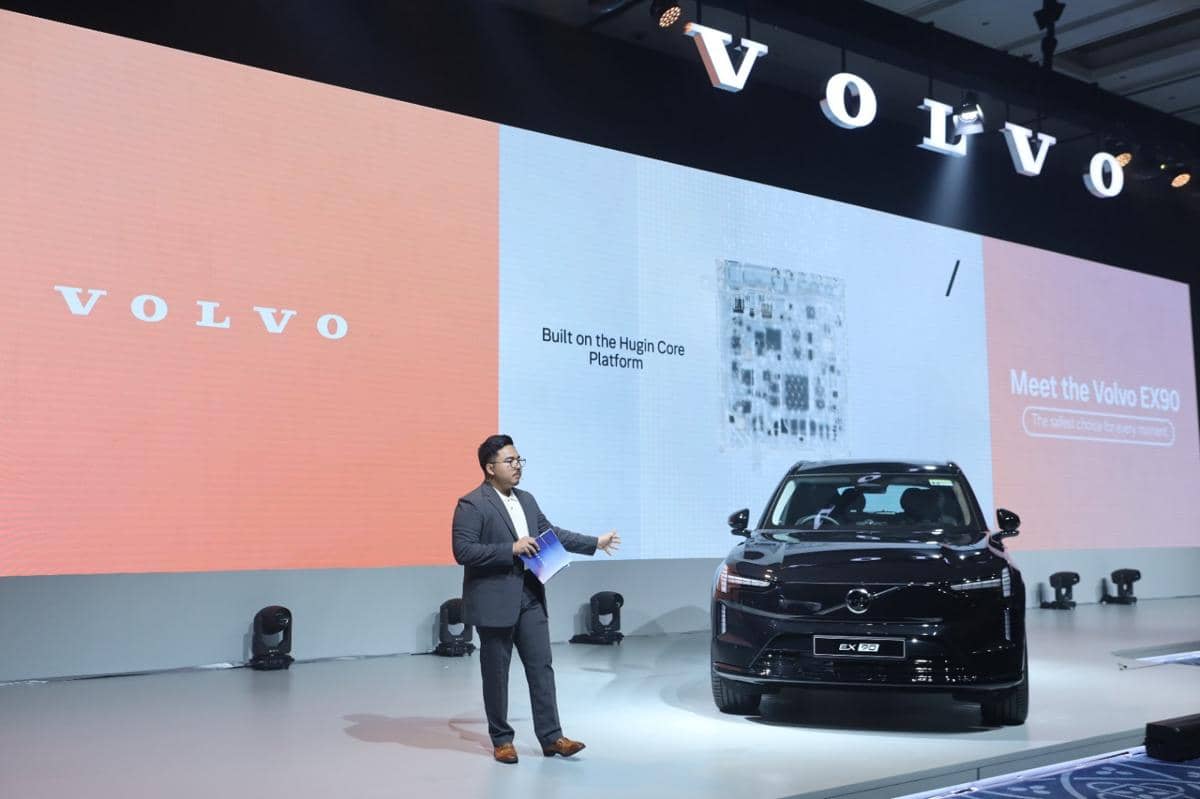 Seorang pembicara berdiri di samping mobil listrik Volvo EX90 hitam saat acara peluncuran di panggung dengan layar besar bertuliskan Volvo.