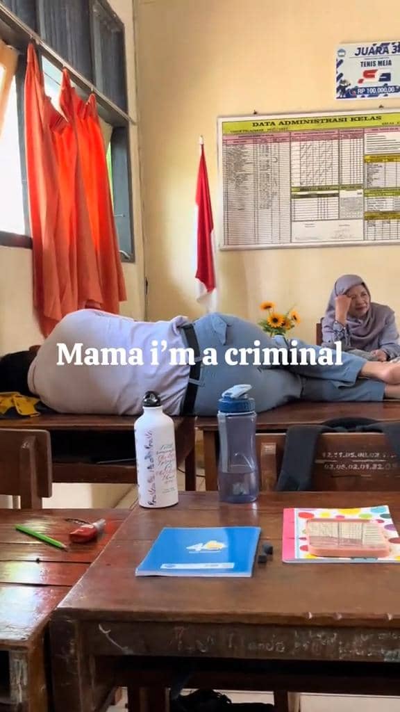 Seorang siswa berbaring di atas meja kelas sementara temannya duduk tersenyum di sebelahnya di dalam ruang kelas sekolah.