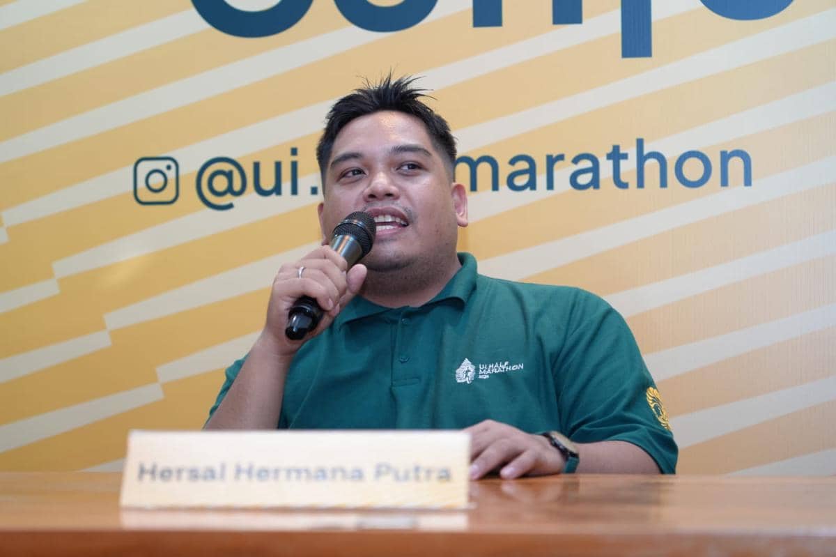 Seorang pria mengenakan kaus hijau berlogo UI Half Marathon berbicara menggunakan mikrofon di depan latar kuning bertulisan media sosial acara.