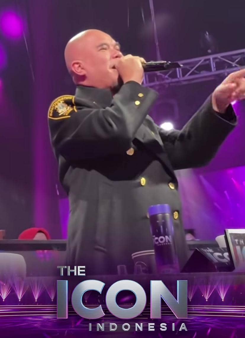 Seorang pria berkepala plontos mengenakan jas hitam dengan kancing emas sedang berbicara menggunakan mikrofon di panggung The Icon Indonesia.