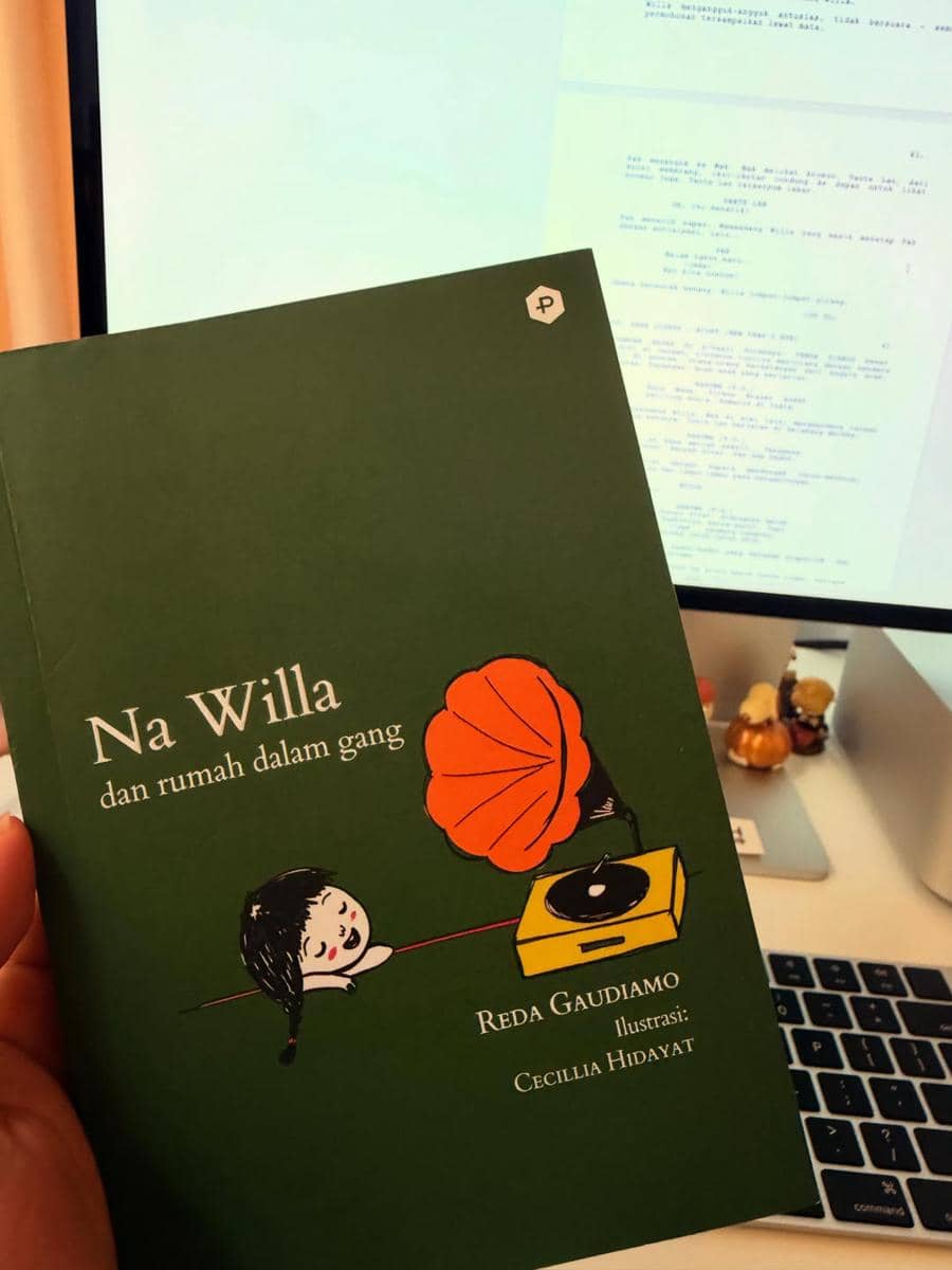 Seseorang memegang buku berjudul Na Willa dan Rumah Dalam Gang karya Reda Gaudiamo dengan ilustrasi Cecilia Hidayat di depan layar komputer.