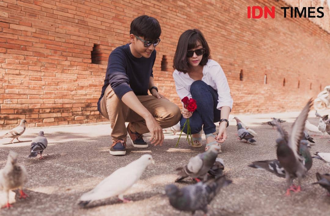 Sepasang anak muda memberi makan burung merpati di area terbuka dengan latar dinding bata merah pada siang hari.