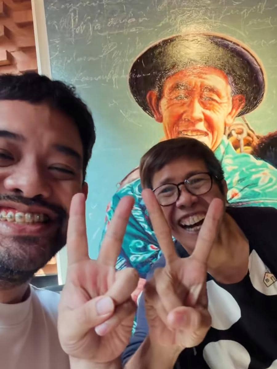 Dua orang tersenyum lebar sambil mengacungkan tanda damai dengan latar belakang mural berwarna cerah bergambar pria tua mengenakan topi.