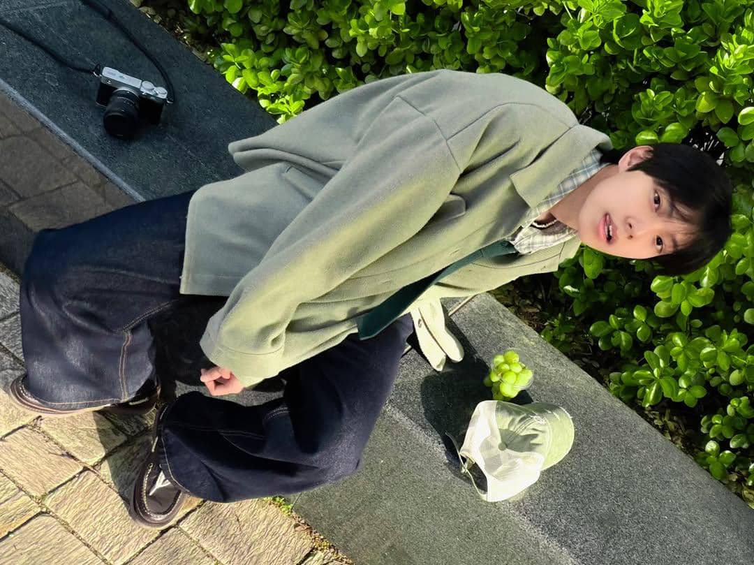 Pria bergaya kasual dengan jaket hijau muda dan celana denim duduk di bangku taman dengan kamera dan anggur hijau di sampingnya.