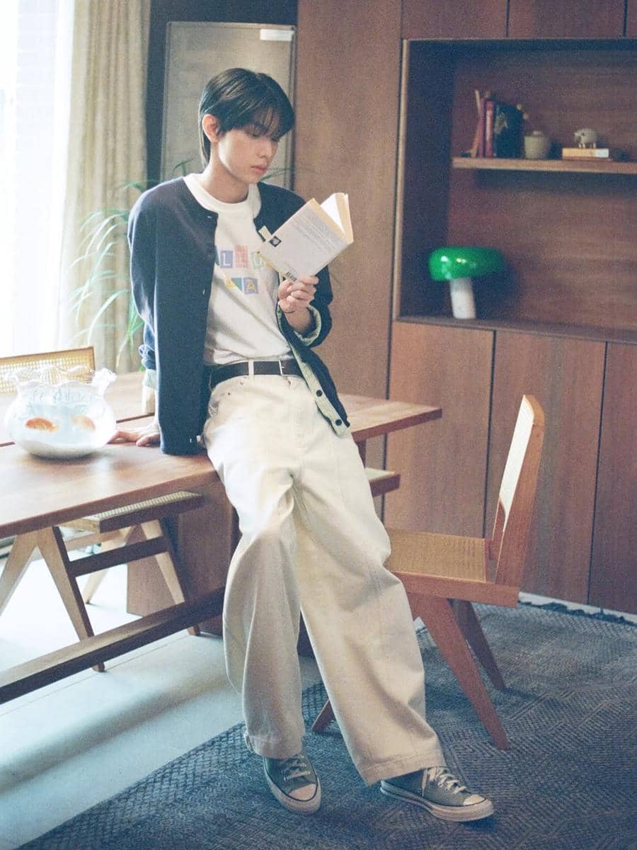 Seorang pria berpenampilan kasual dengan gaya soft boy mengenakan kardigan gelap, kaus putih, dan celana panjang krem sambil membaca buku di ruang tamu.