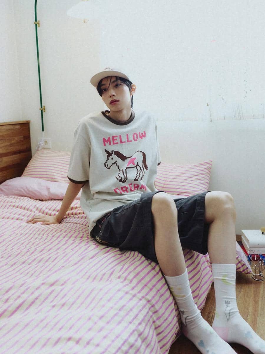 Seorang pria bergaya kasual duduk di atas tempat tidur dengan seprai bergaris pink, mengenakan kaus bergambar kuda, celana pendek hitam, dan topi.