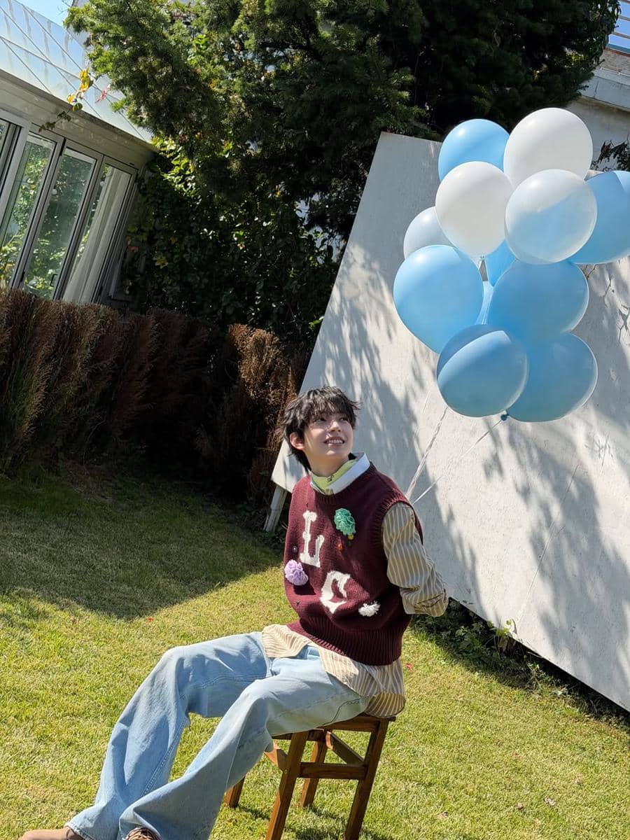 Seorang pria bergaya soft boy duduk di kursi taman sambil memegang balon biru dan putih di bawah sinar matahari.