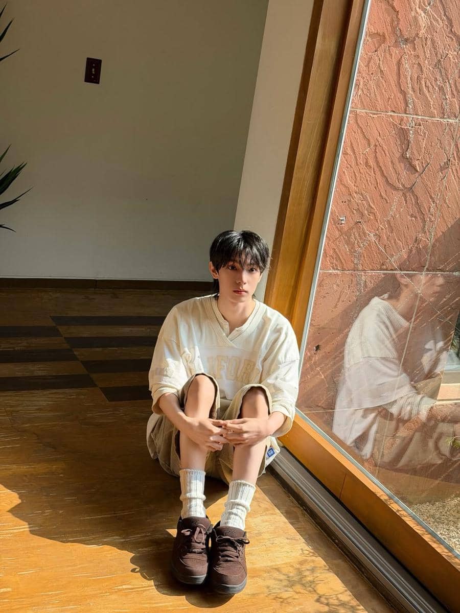 Seorang pria bergaya soft boy duduk di lantai dekat jendela besar dengan cahaya matahari masuk, mengenakan sweater krem, celana pendek, dan sepatu cokelat.