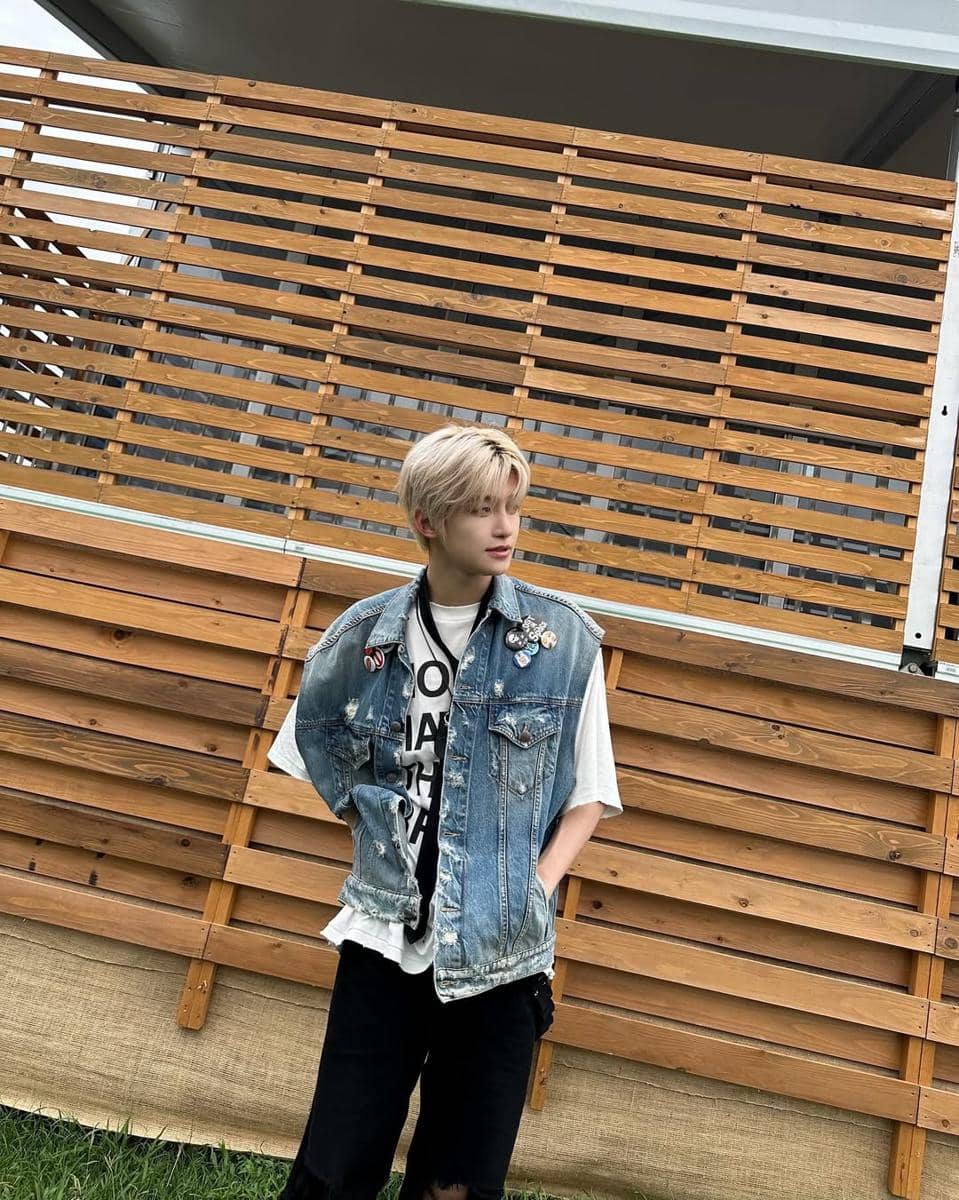 Seorang pria berambut pirang mengenakan kaus putih dan rompi denim dengan pin bergaya street style berdiri di depan pagar kayu.