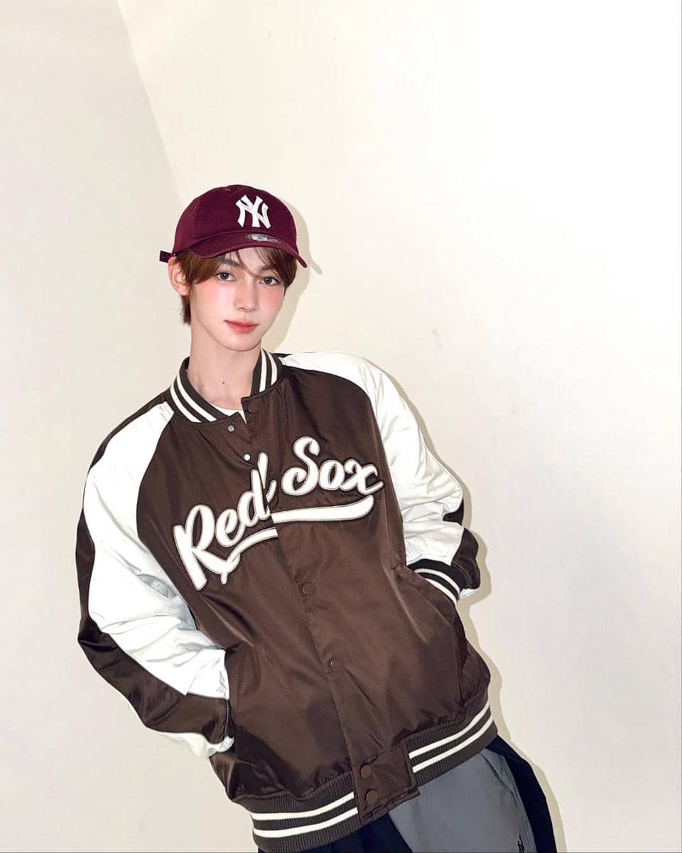 Seorang pria mengenakan jaket varsity cokelat bertuliskan Red Sox dan topi baseball merah marun dengan logo NY di depan latar polos.