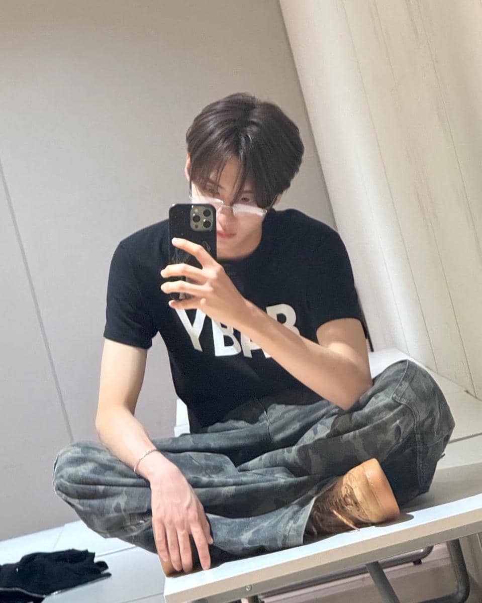 Seorang pria berpose duduk di atas meja sambil berfoto di cermin, mengenakan kaus hitam bertulisan YBAB, celana motif kamuflase, dan sepatu cokelat.