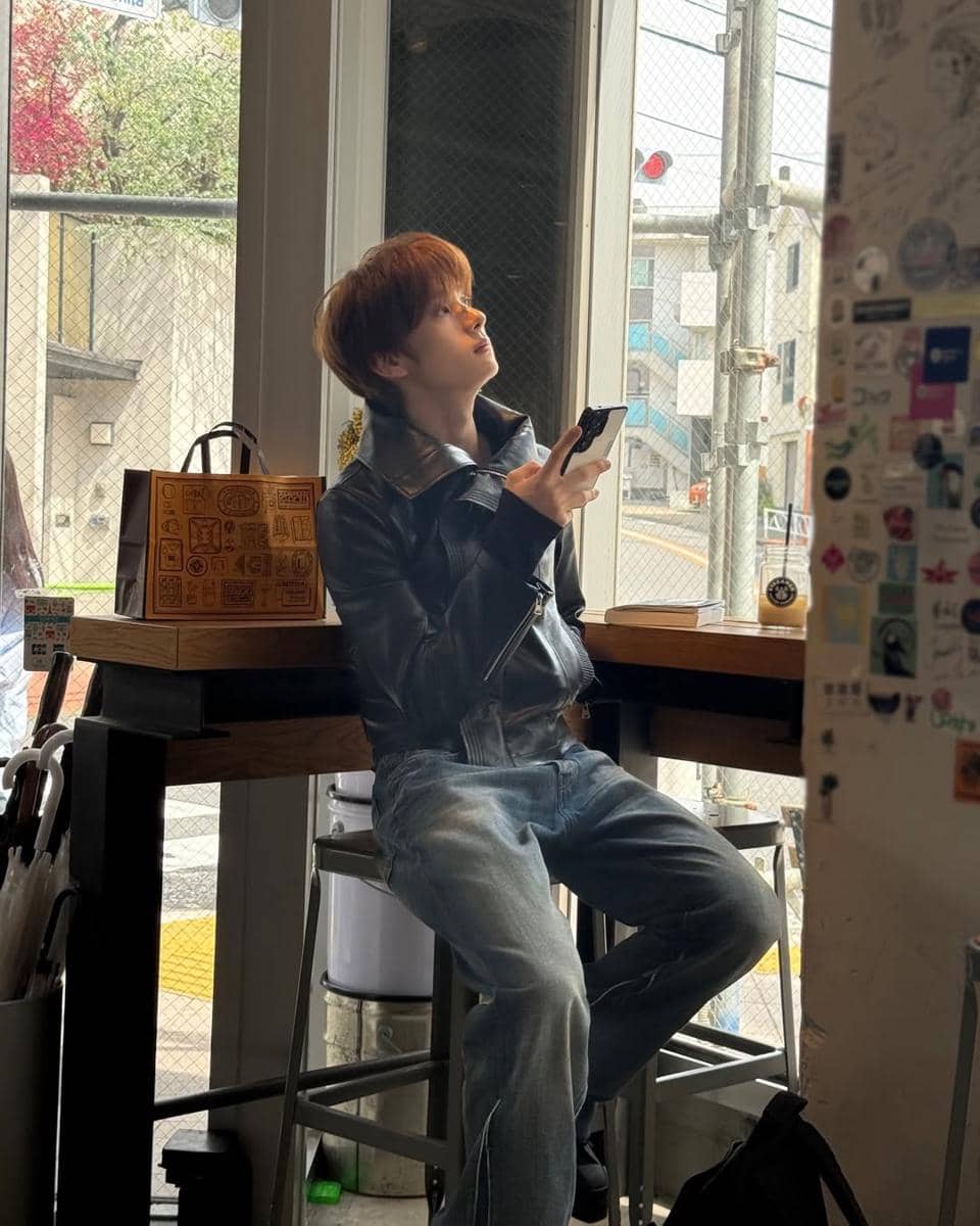 Seorang pria berjaket kulit hitam dan celana jeans duduk di kursi tinggi dekat jendela sambil memegang ponsel di sebuah kafe.