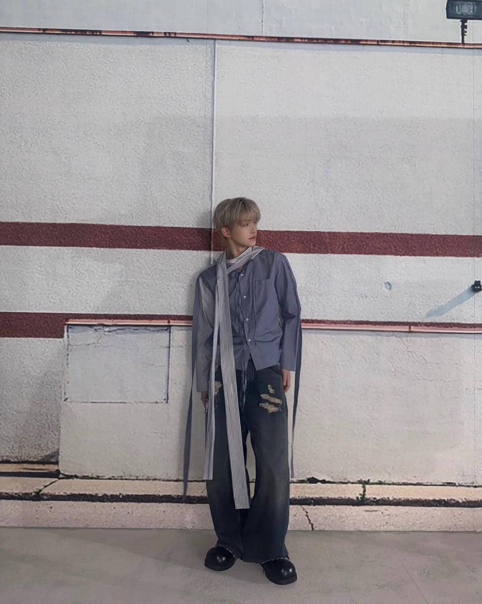 Seorang pria berpose dengan gaya street style mengenakan kemeja abu-abu longgar, celana jeans lebar robek, dan sepatu hitam di depan dinding bergaris merah.