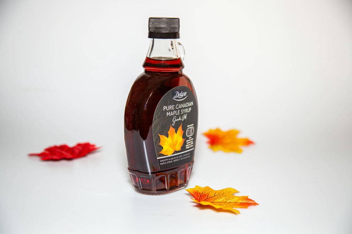 Botol sirup maple murni asal Kanada dengan label hitam berdiri di atas permukaan putih, dikelilingi daun maple berwarna merah dan kuning.