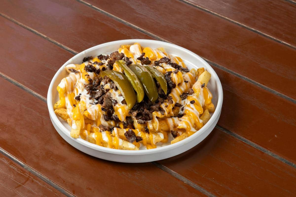 Semangkuk poutine berisi kentang goreng dengan saus keju leleh, potongan daging, saus putih, dan irisan acar hijau di atas meja kayu.