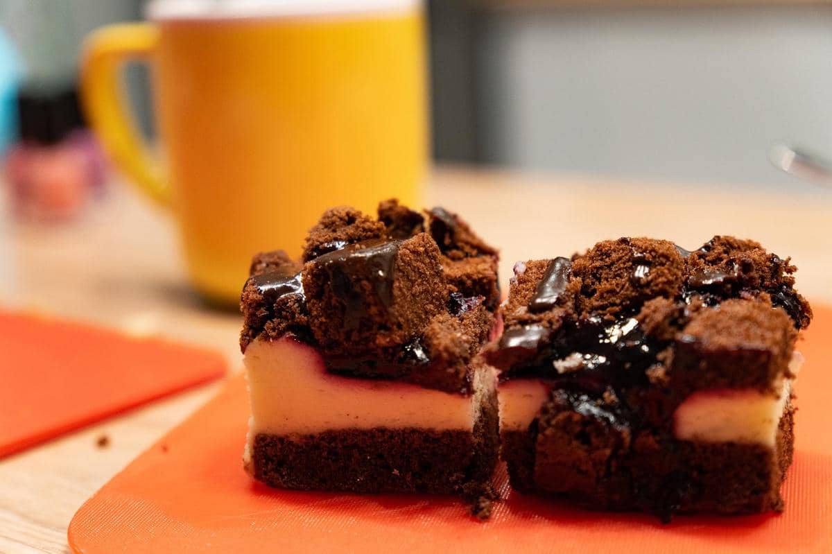 Dua potong nanaimo bars berlapis cokelat, krim, dan remah kue disajikan di atas alas oranye dengan cangkir kuning di latar belakang.