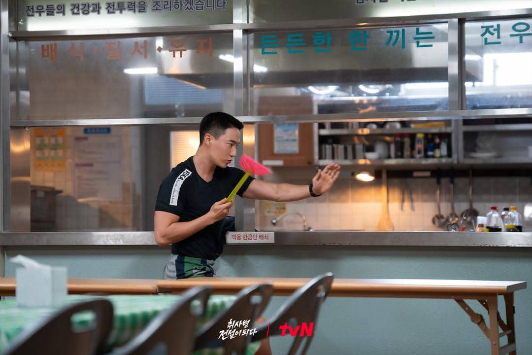 Seorang pria berpakaian olahraga memegang pemukul lalat berwarna merah muda di dapur kantin militer dalam adegan drama The Legend of Kitchen Soldier.