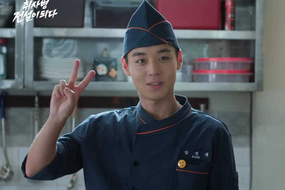 Seorang pria muda mengenakan seragam koki berwarna biru tua dengan topi dapur, tersenyum sambil mengangkat tangan membentuk tanda V di dapur.
