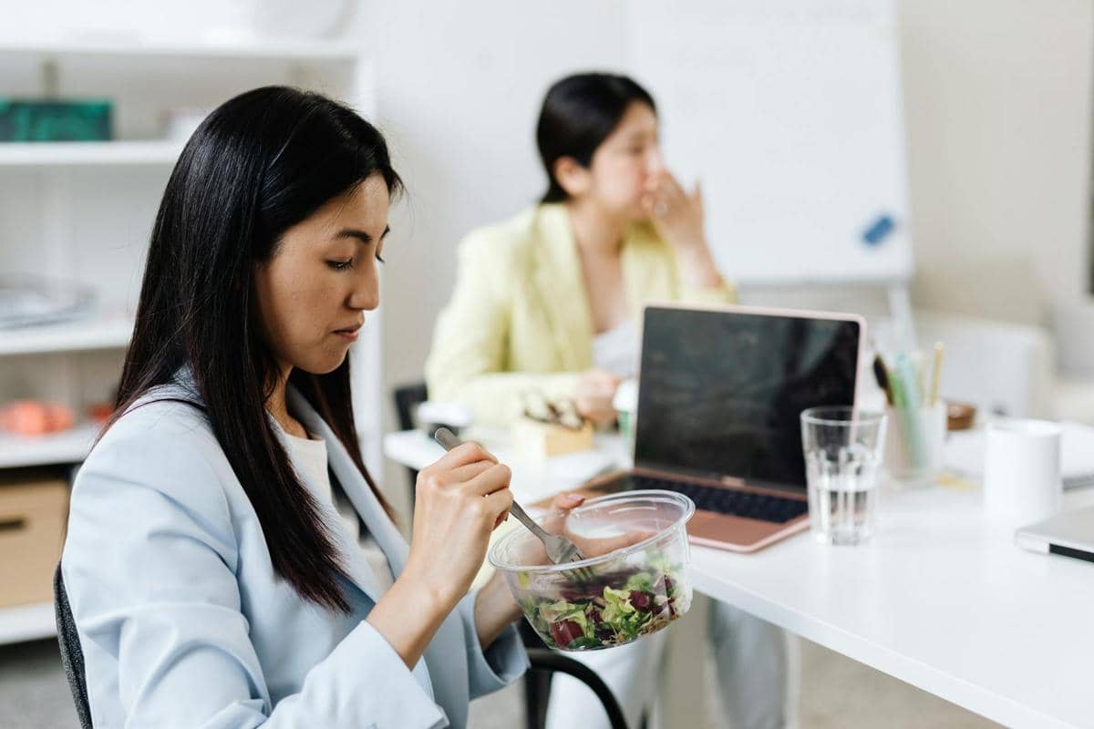 Seorang pekerja kantoran mengenakan blazer biru muda sedang makan salad di meja kerja dengan laptop dan gelas air di depannya.