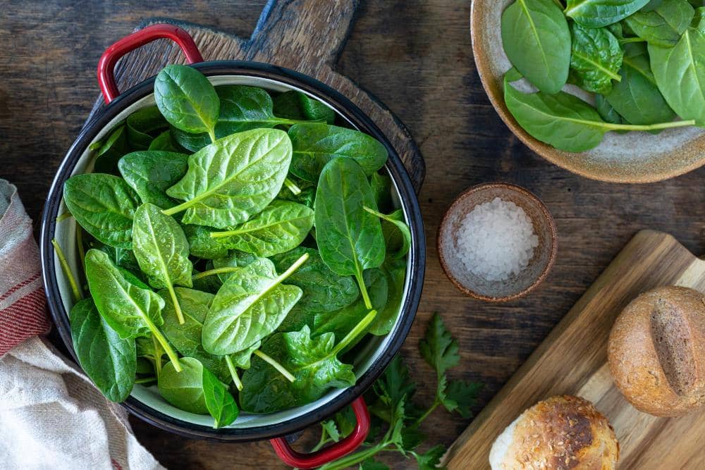 Daun bayam segar berwarna hijau ditempatkan dalam panci logam di atas meja kayu bersama garam dan roti di sekitarnya.