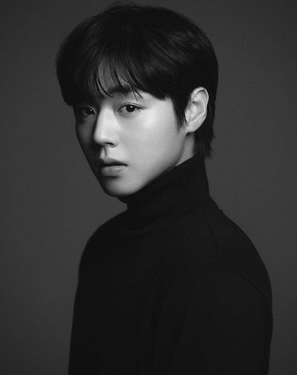 Potret seorang pria berambut hitam mengenakan sweater berleher tinggi hitam dalam foto hitam putih dengan latar polos.