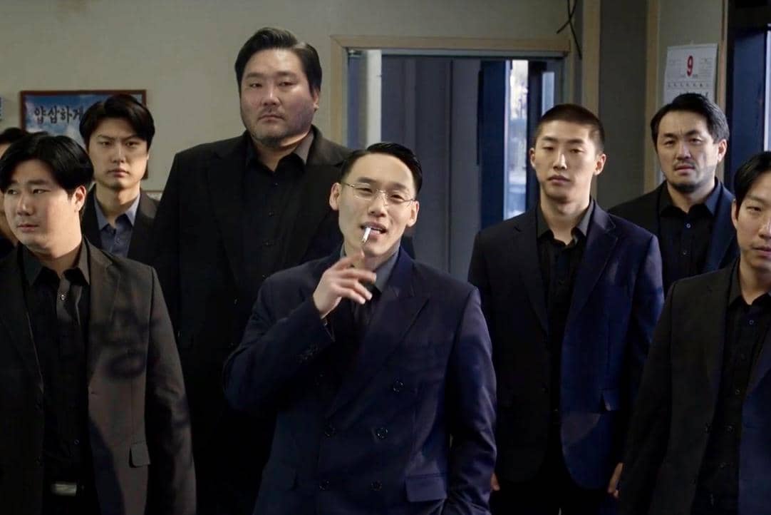 Beberapa pria berpakaian jas hitam berdiri di dalam ruangan, salah satunya memegang rokok dalam cuplikan drama Korea Gold Land.