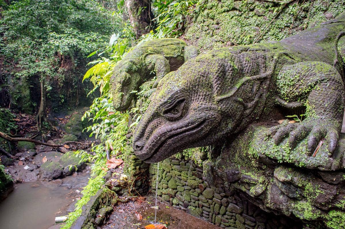 Patung komodo berlumut di Hutan Monyet Ubud, Bali, dengan latar pepohonan hijau dan aliran air kecil di sekitarnya.