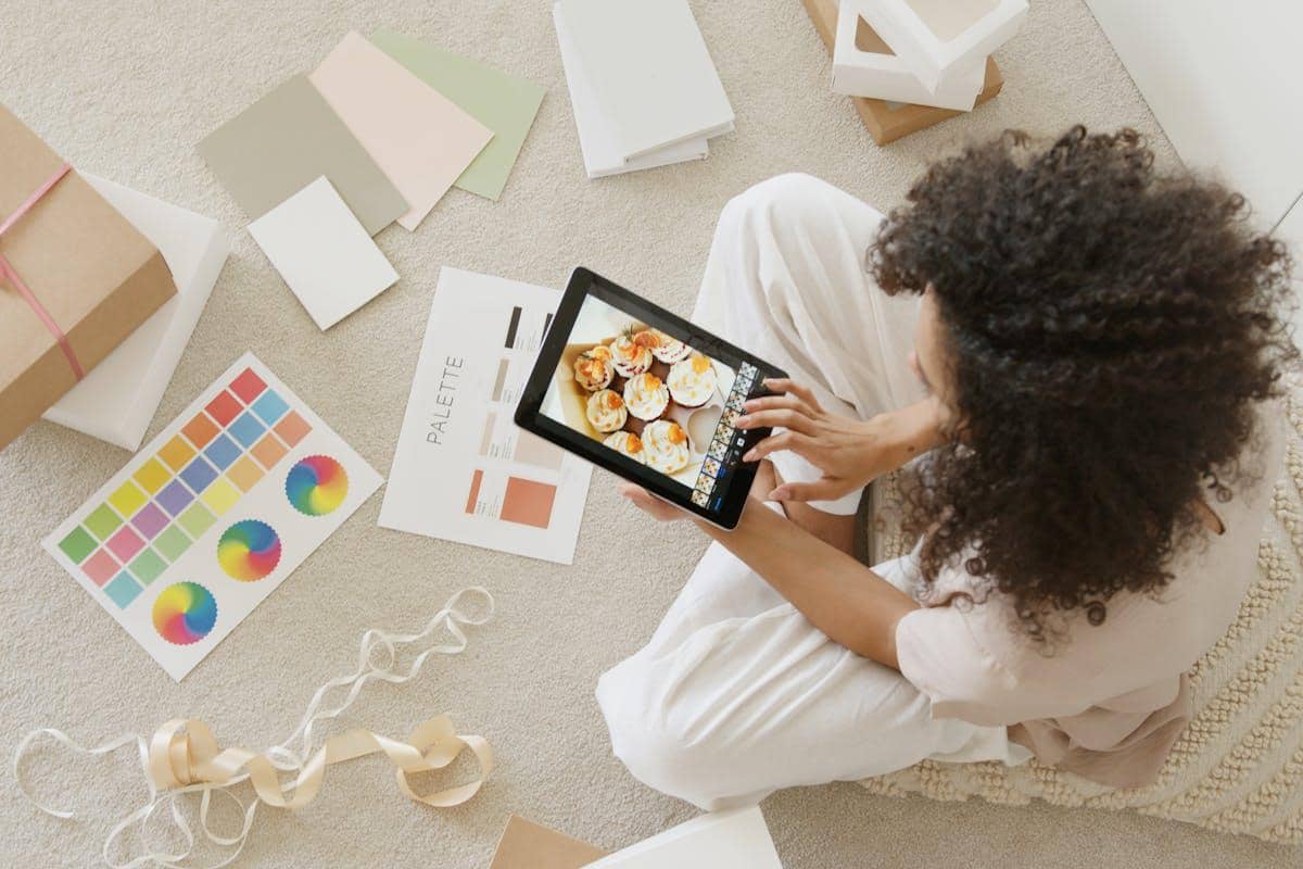 Seorang perempuan duduk di lantai sambil mengedit foto kue di tablet, dikelilingi palet warna, pita, dan bahan desain produk.