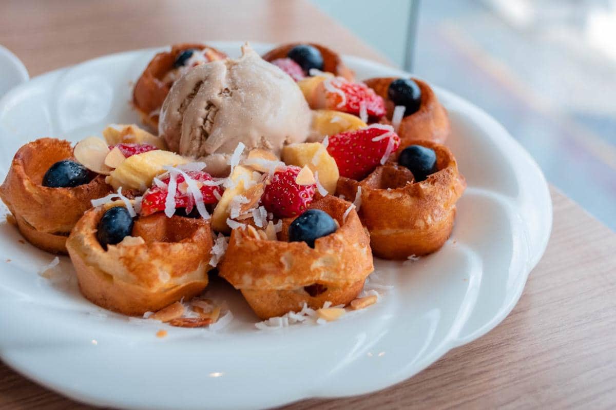 Sepiring es krim goreng disajikan dengan potongan stroberi, blueberry, pisang, dan taburan kelapa parut di atas piring putih.