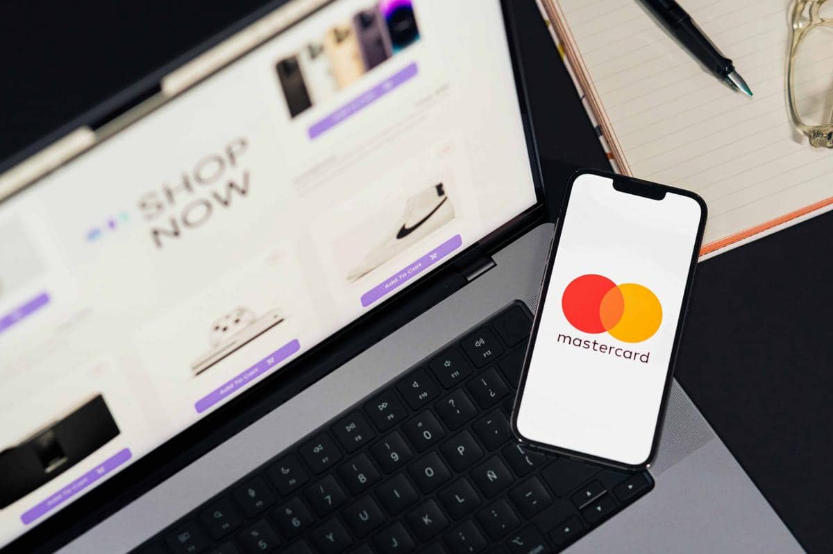 Ponsel menampilkan logo Mastercard di atas laptop yang membuka situs belanja online dengan tulisan Shop Now di layar.