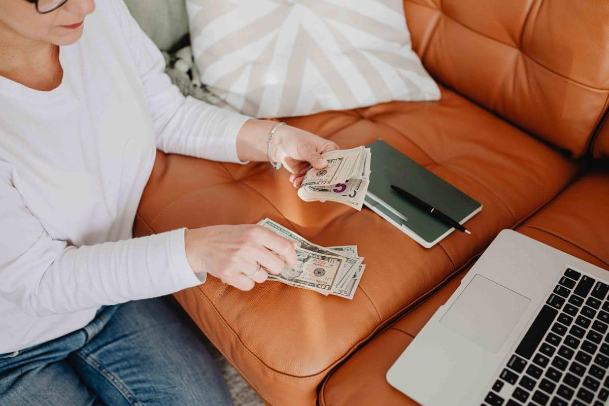 Seseorang mengenakan pakaian kasual sedang menghitung lembaran uang di atas sofa kulit dengan laptop dan buku catatan di sampingnya.