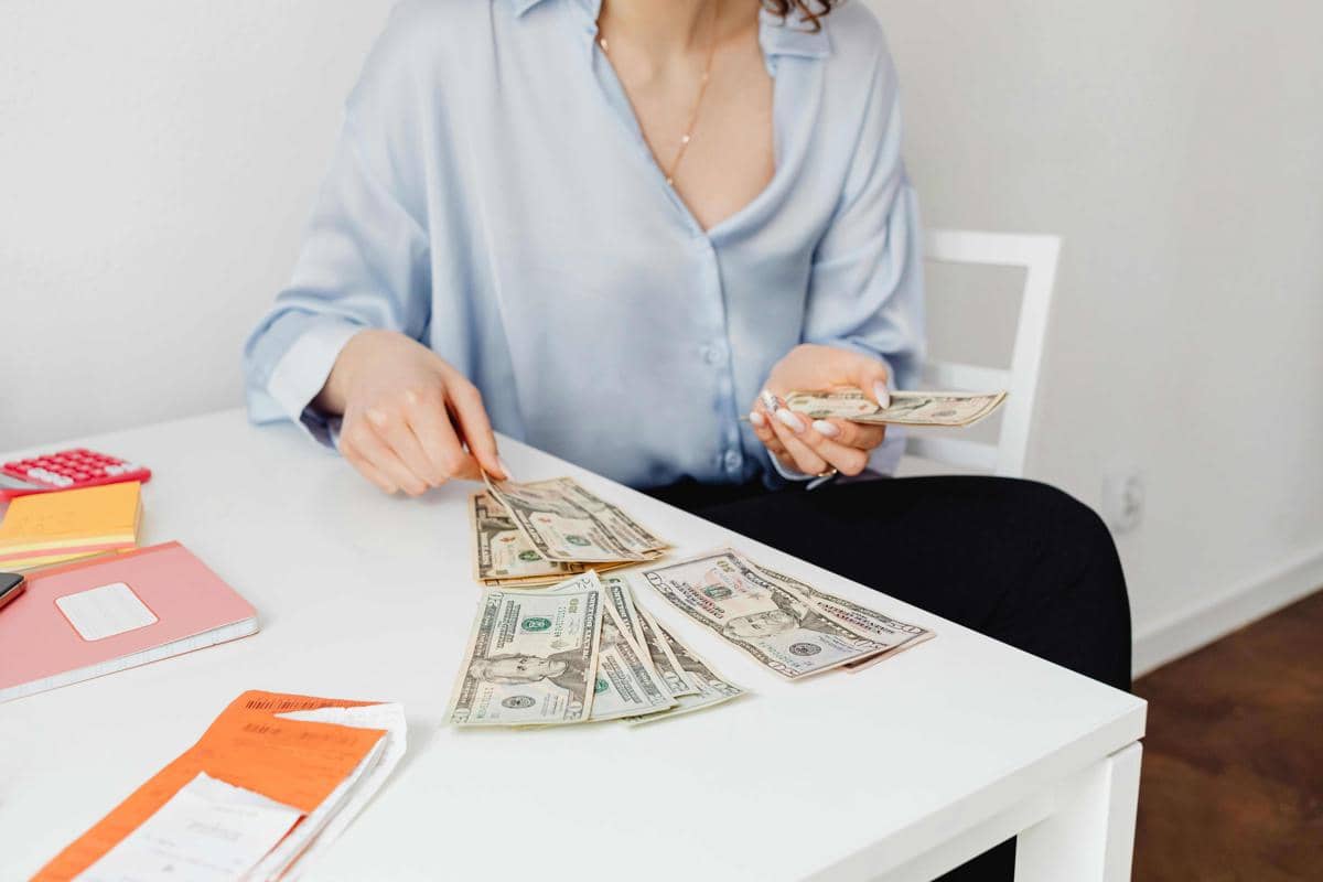 Seorang wanita mengenakan kemeja biru muda menghitung dan menyusun lembaran uang dolar di atas meja putih dengan buku catatan dan kalkulator.
