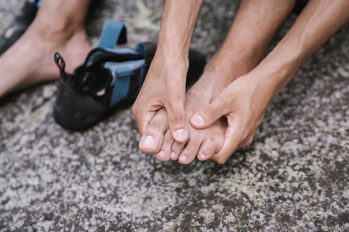 Seseorang memegang jari kaki yang tampak nyeri di atas permukaan berbatu dengan sepatu dilepaskan di sampingnya.