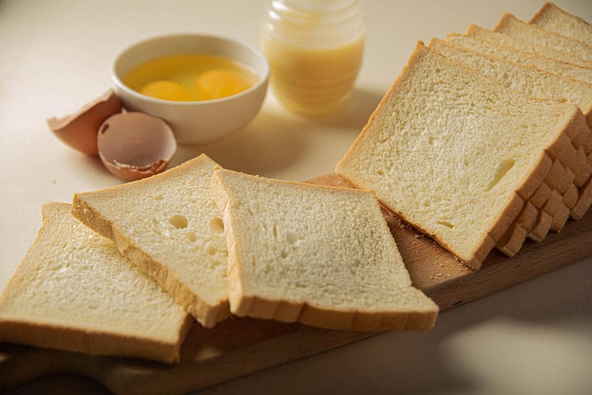 Beberapa lembar roti tawar tersusun di atas talenan kayu dengan latar mangkuk berisi telur mentah dan cangkang telur di meja.