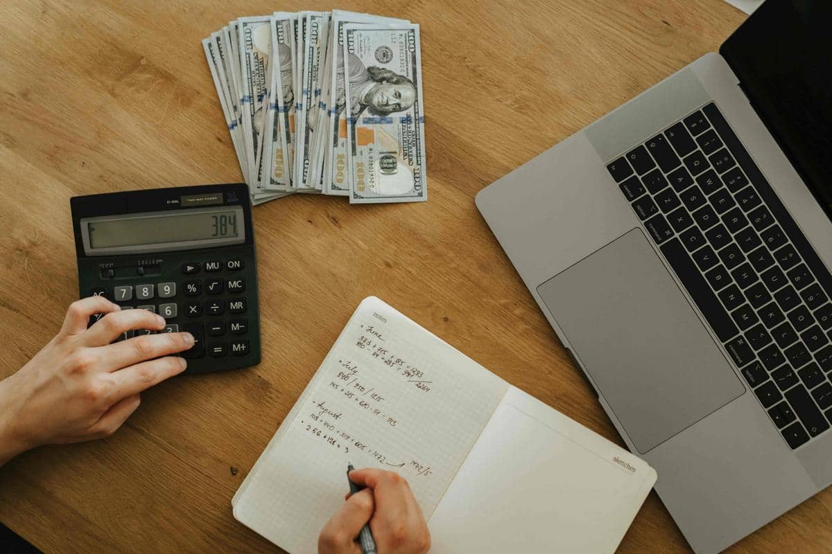 Seseorang menghitung anggaran dengan kalkulator di meja kayu, disertai uang dolar, buku catatan, dan laptop terbuka di sampingnya.