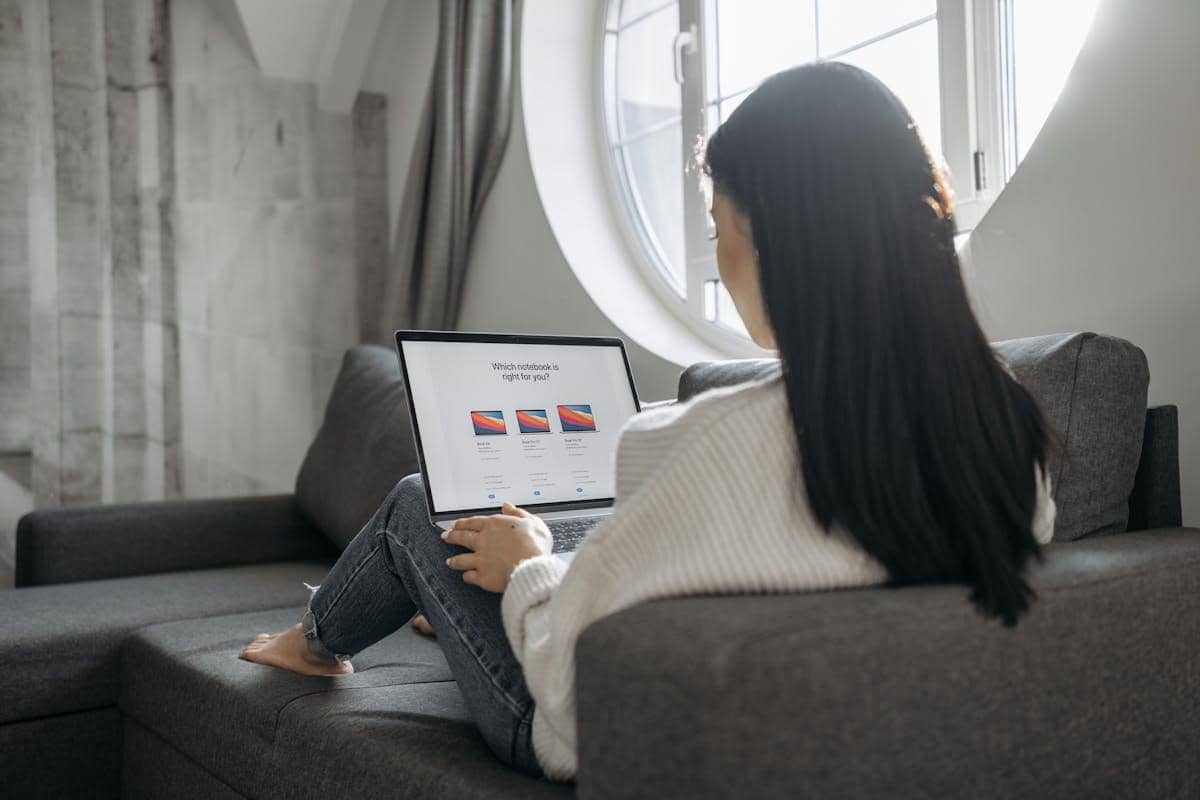 Seorang wanita duduk di sofa sambil menggunakan laptop untuk mengisi survei online di ruangan dengan jendela bundar.
