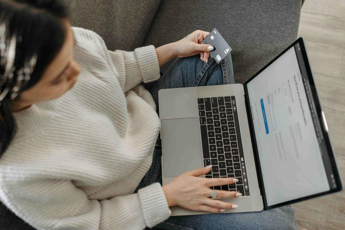 Seseorang duduk di sofa sambil memegang kartu kredit dan menggunakan laptop untuk berbelanja daring di rumah.