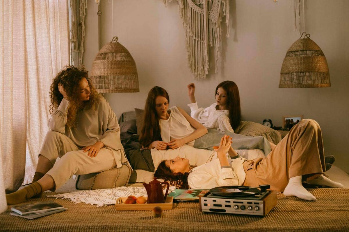Empat perempuan bersantai di kamar bernuansa hangat dengan dekorasi bohemian, berbincang sambil mendengarkan musik dari pemutar piringan hitam.