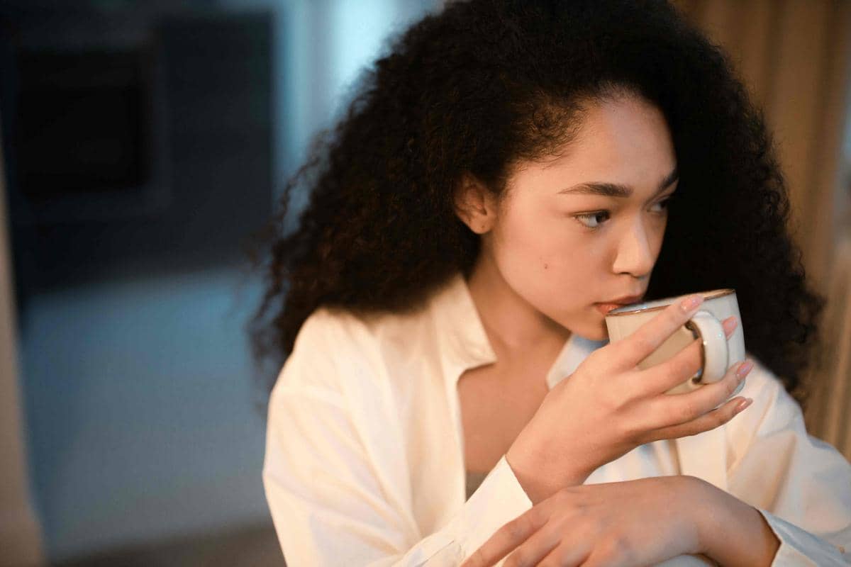 Seorang perempuan berambut keriting mengenakan kemeja putih sedang menikmati secangkir kopi di dalam ruangan dengan pencahayaan hangat.