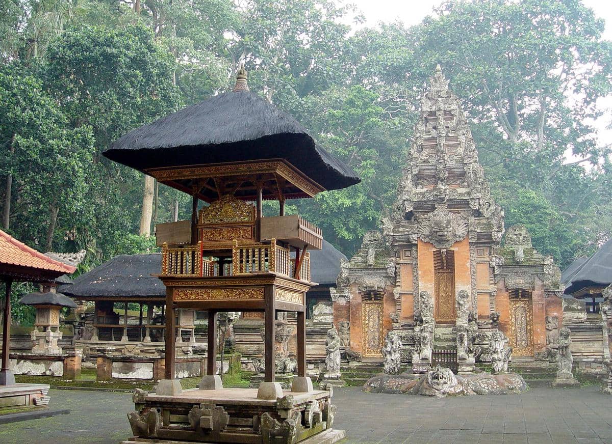 Arsitektur tradisional Pura Dalem Agung di Hutan Monyet Ubud dengan ukiran batu dan atap jerami di tengah pepohonan hijau.