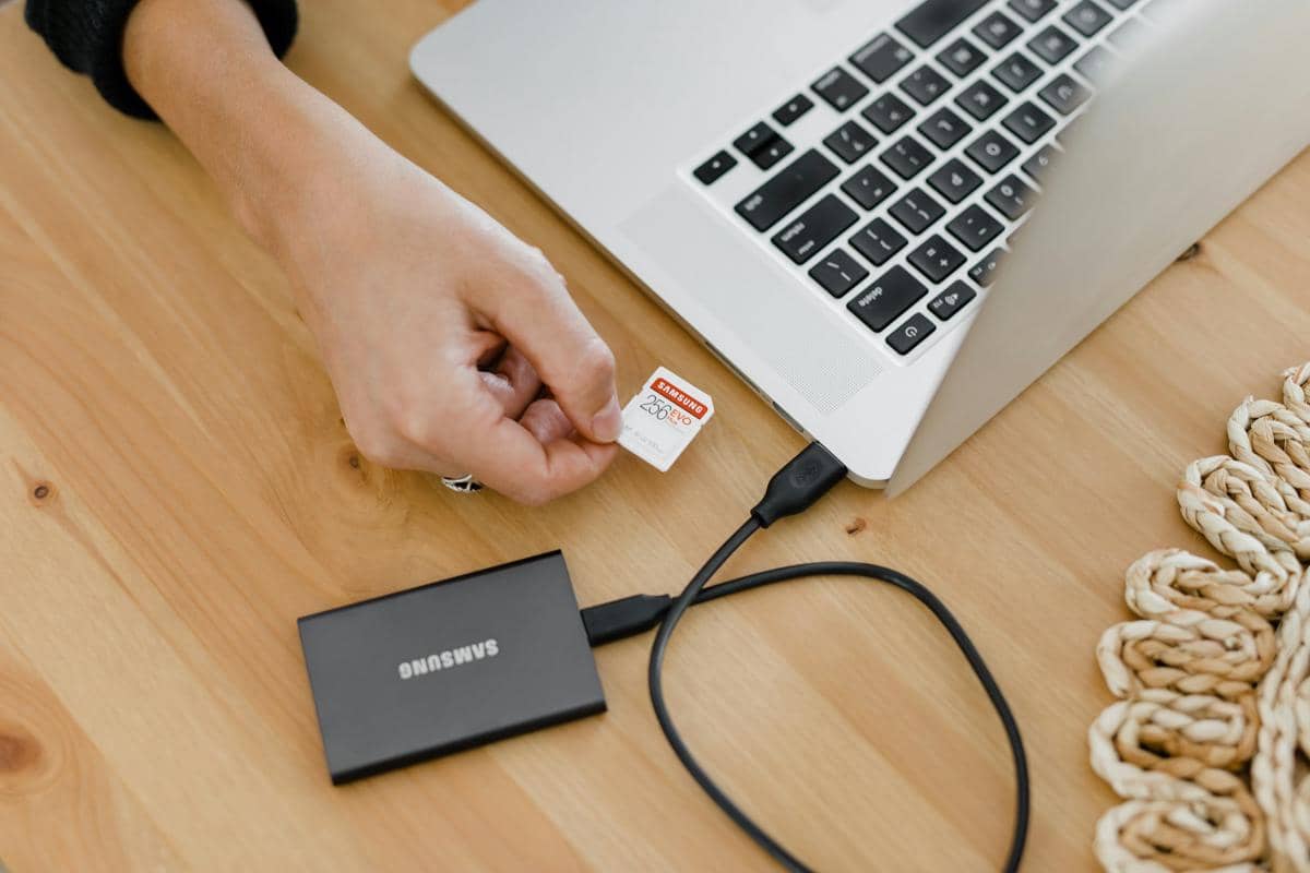 Tangan memasukkan kartu memori ke laptop yang terhubung dengan SSD eksternal Samsung di atas meja kayu.