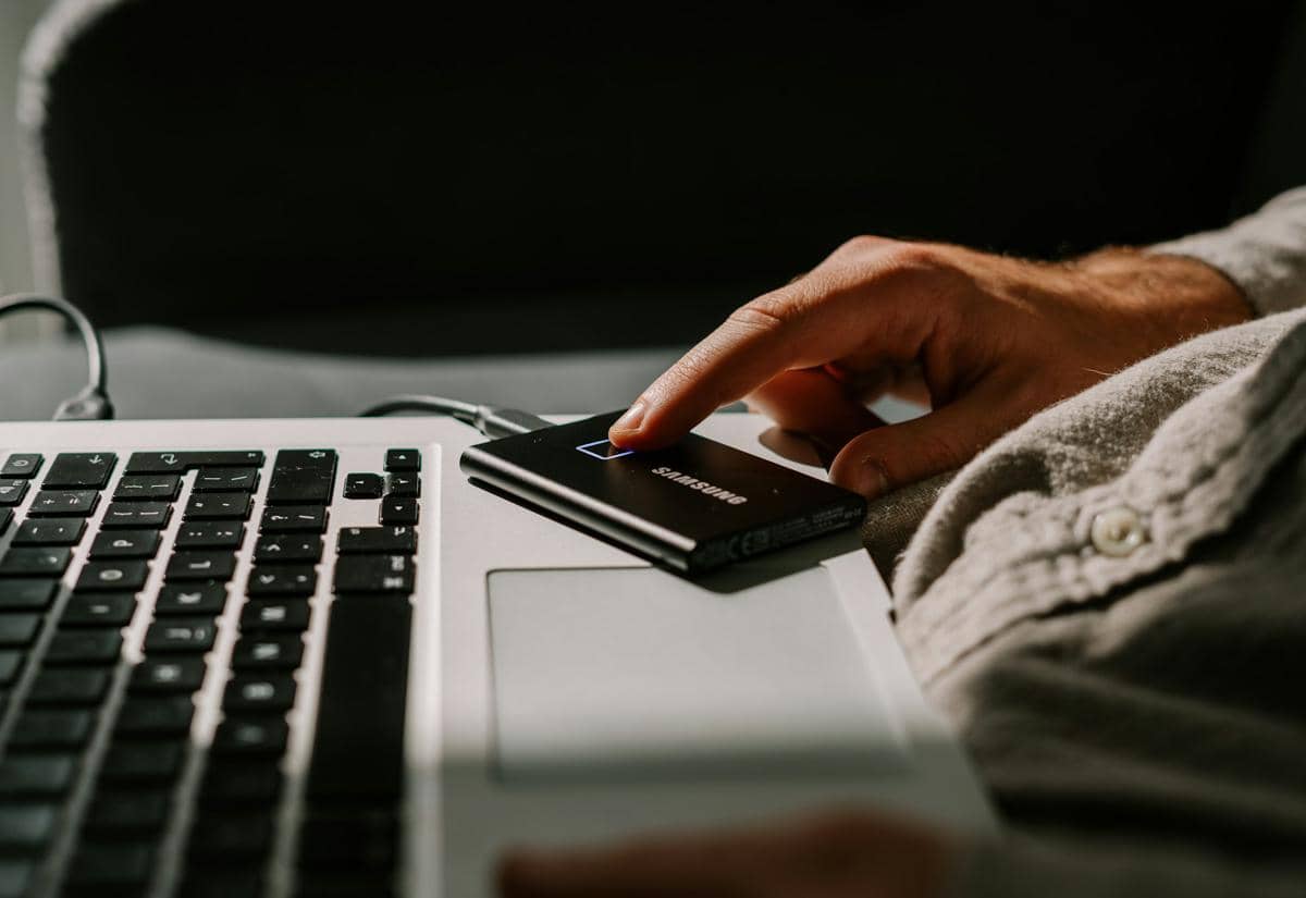 Seseorang menyentuh SSD eksternal Samsung yang terhubung ke laptop berwarna perak di atas meja kerja dengan pencahayaan lembut.