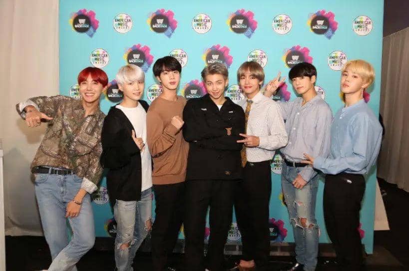 Tujuh anggota grup musik Korea berpose bersama di depan latar belakang bertema American Music Awards 2017 dengan ekspresi ceria.