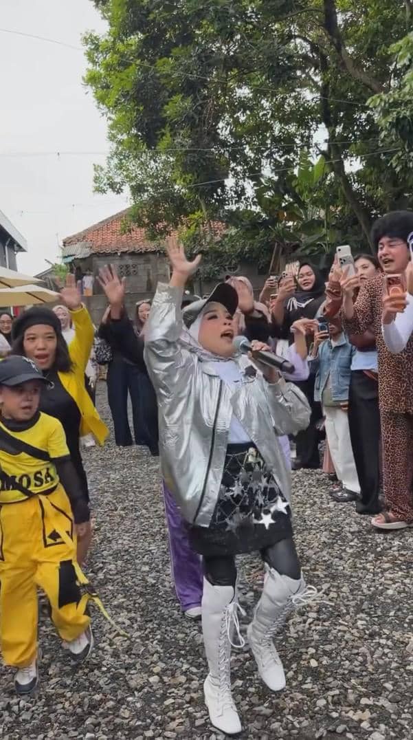 Seorang perempuan bernyanyi dengan mikrofon mengenakan jaket perak dan sepatu putih di pesta ulang tahun outdoor, dikelilingi tamu yang bersorak.