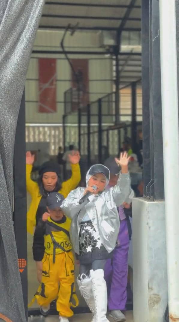 Seorang anak mengenakan jaket perak dan sepatu bot putih bernyanyi dengan mikrofon, diikuti beberapa anak lain dalam kostum warna-warni.