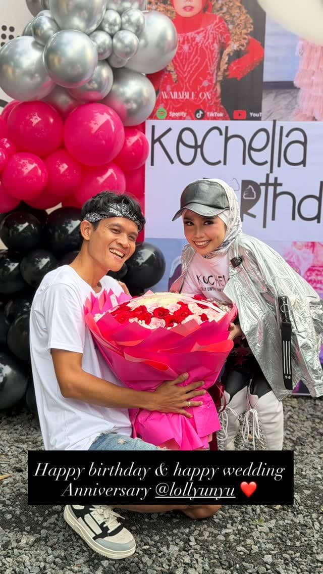 Seorang pria dan wanita tersenyum sambil memegang buket bunga besar berwarna merah muda di acara ulang tahun bertema balon merah, hitam, dan perak.