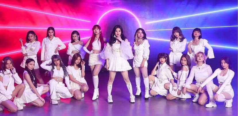 Grup idola perempuan MNL48 berpose bersama mengenakan busana putih dengan latar belakang cahaya neon ungu dan merah muda.
