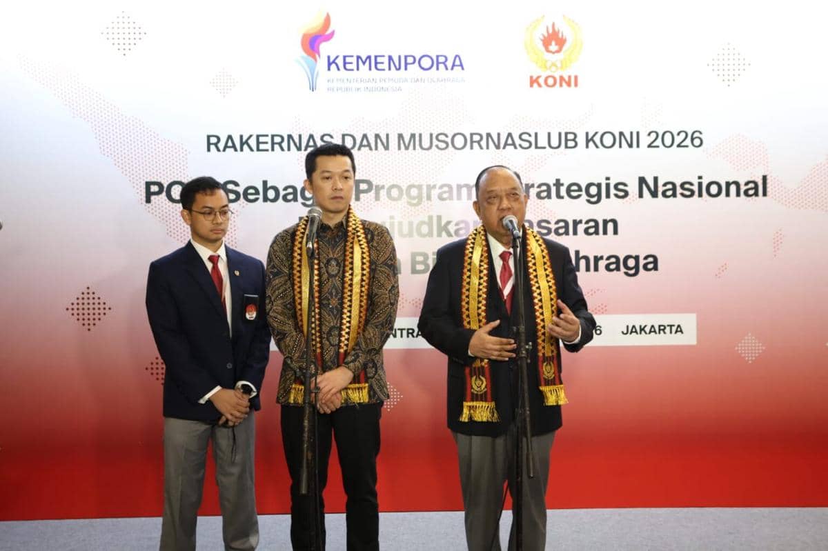 Tiga pria mengenakan pakaian formal dan selendang tradisional berbicara di podium saat Rakernas dan Musornaslub KONI 2026 di Jakarta.
