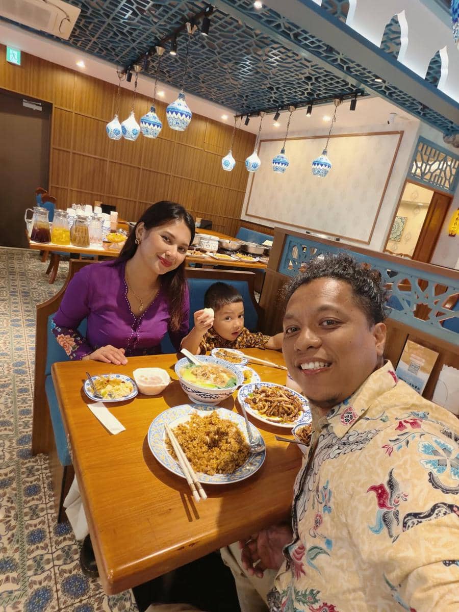 Pasangan dan anak kecil mengenakan batik dan kebaya sedang makan bersama di restoran bergaya Jepang dengan dekorasi biru putih.