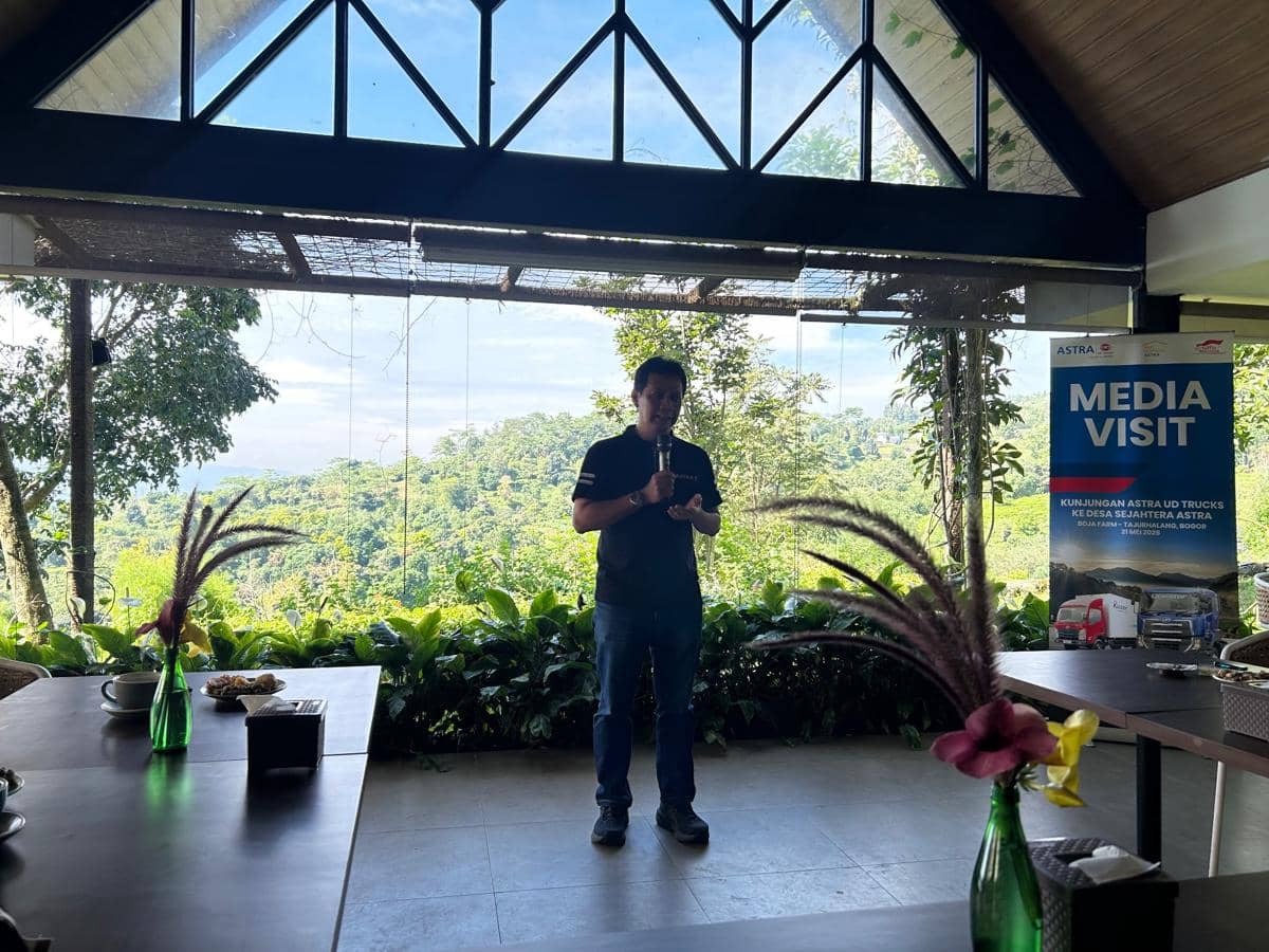 Seorang pria berbicara menggunakan mikrofon di depan ruangan dengan pemandangan alam hijau saat acara media visit Astra UD Trucks di Bogor.